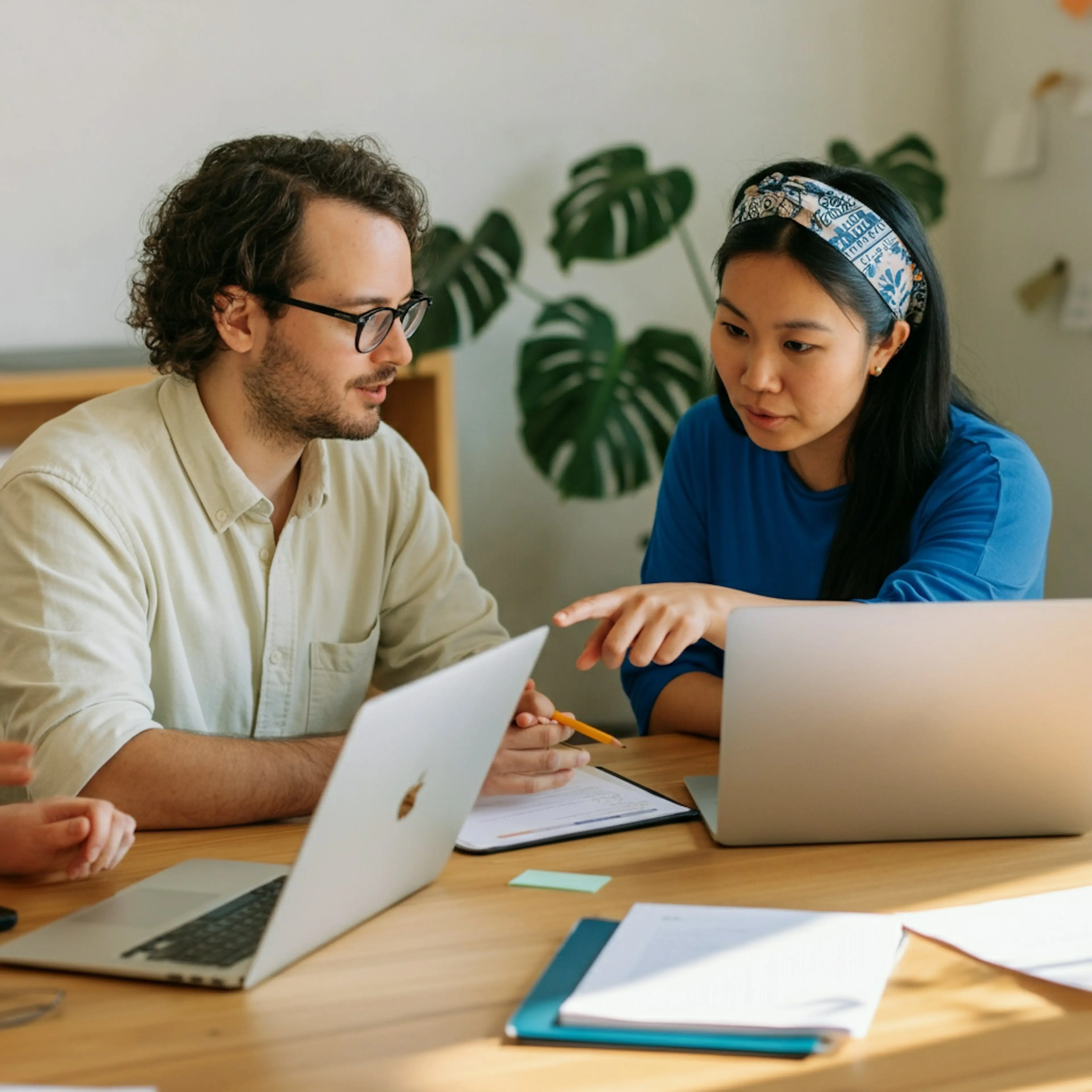 A man, a woman, and a third person who is out of picture are sitting at a desk with their laptops and a notepad. They are discussing something and the woman is pointing towards the third person's laptop.