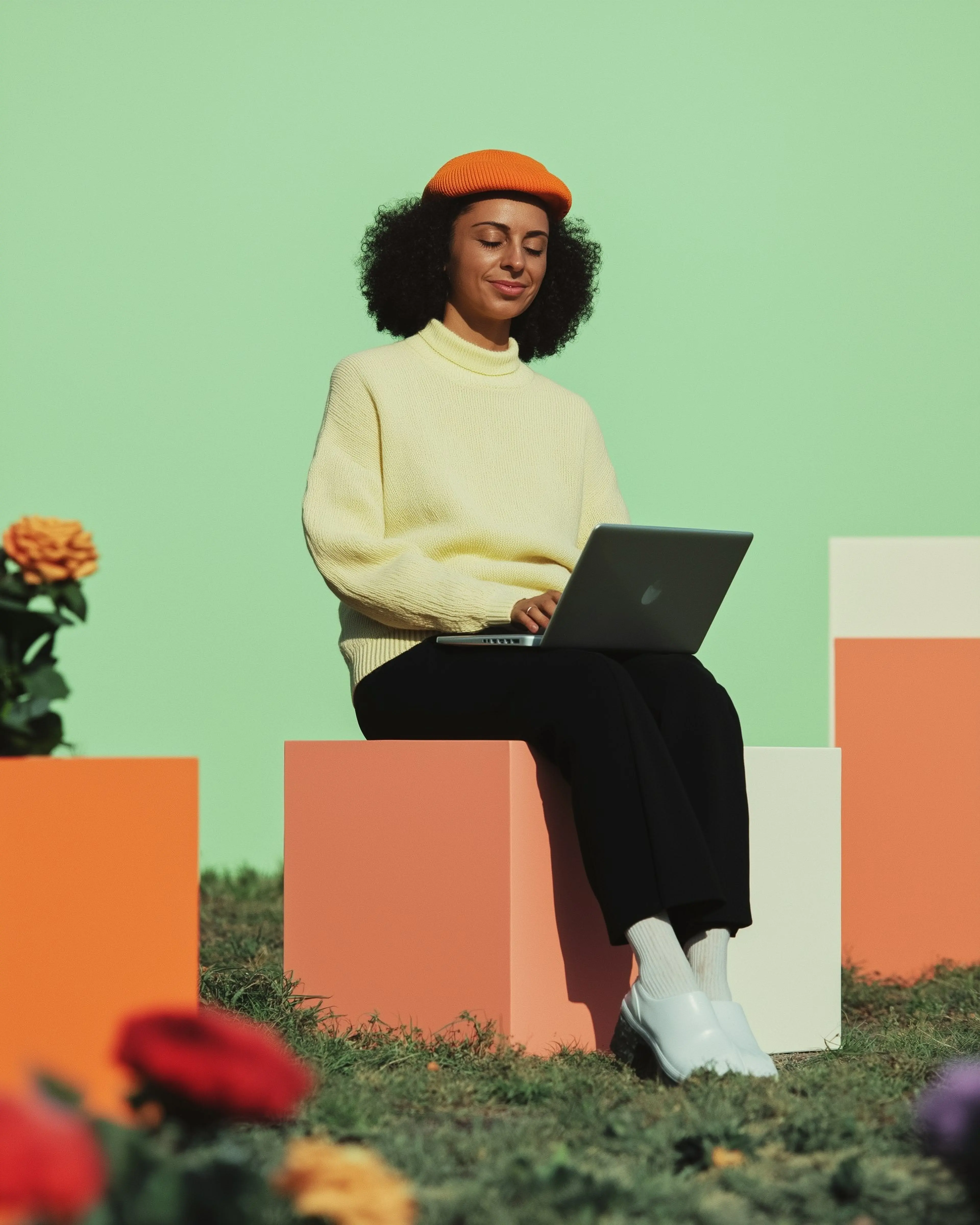 A woman it on a cube in a green screen studio with many other cubes around her. The ground is covered by artificial moss and rose bushes. She has a laptop in her lap, with both hand on the keyboard, and is looking down.