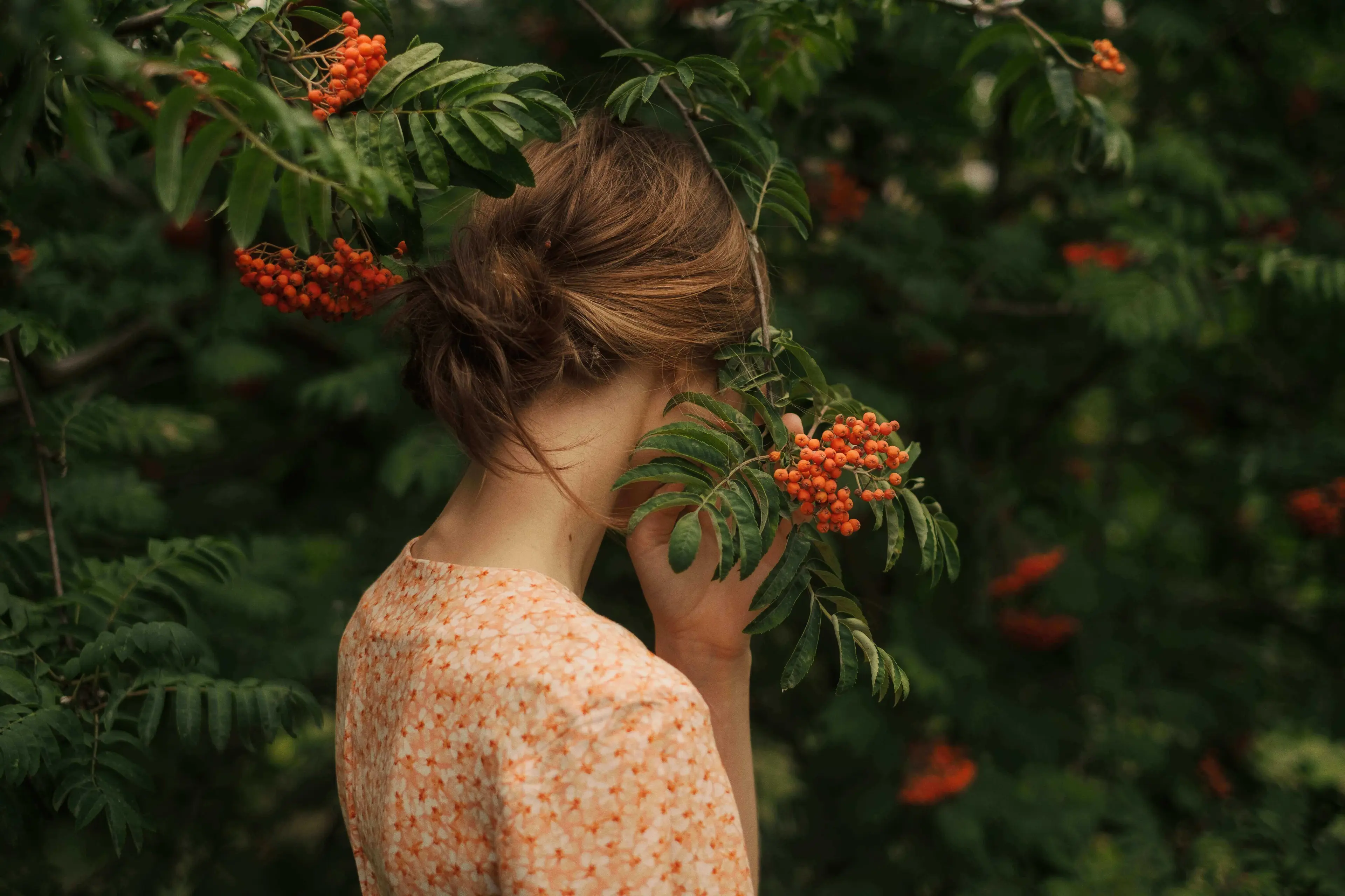 A person with brown hair stands amid lush greenery and vibrant orange berries.