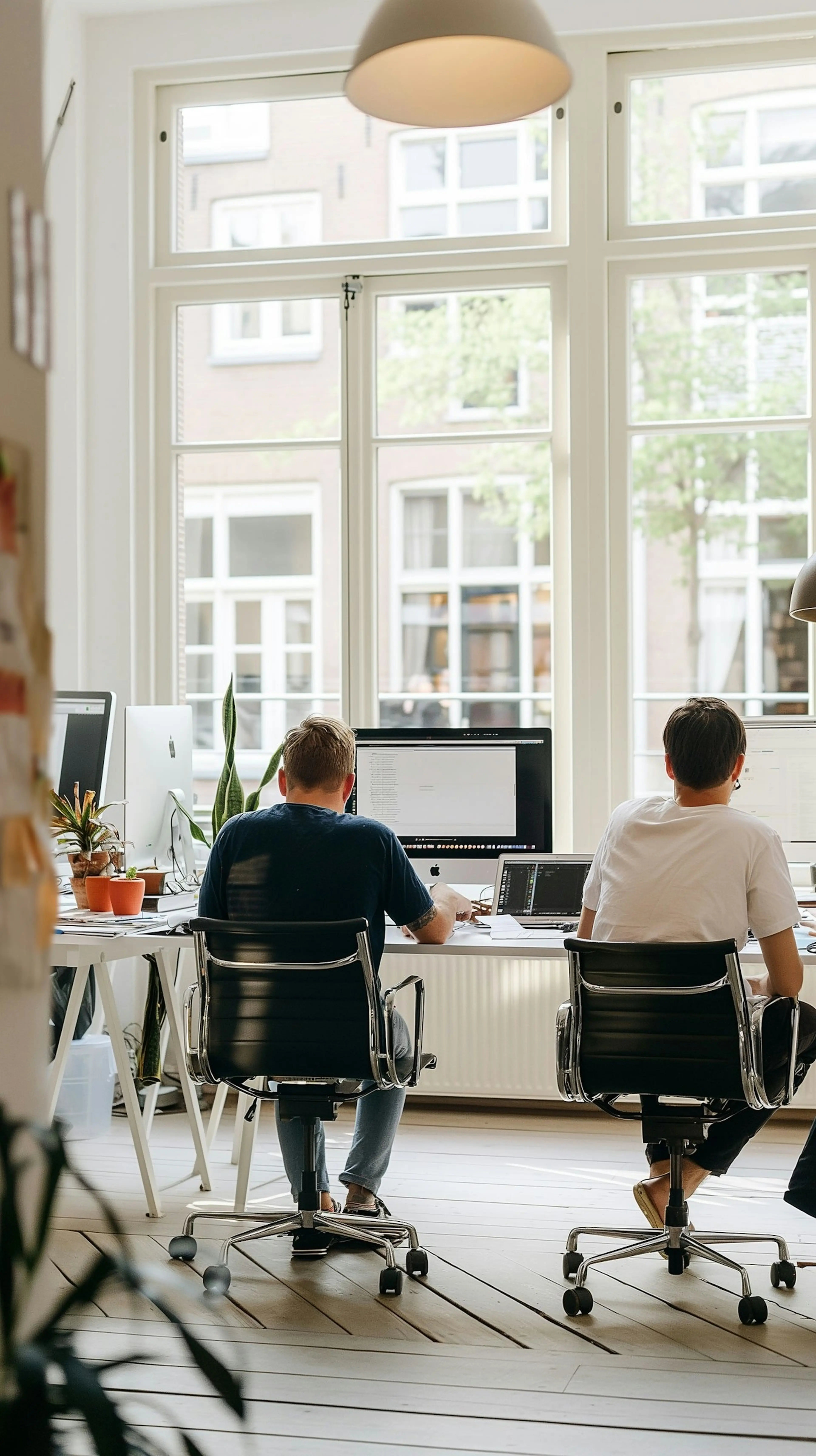 Two men sitting next to each other, each in front of a monitor. The table is positioned in front of a window and the weather outside is sunny.